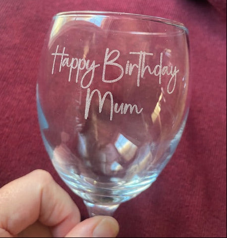 Clear glass wine glass with 'Happy Birthday Mum' engraving held by a person wearing a red sweater.