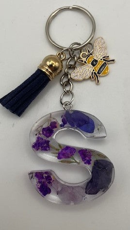 A personalized resin letter 'S' keychain featuring dried purple flowers, a small gold and black bee charm, and a navy blue tassel with a gold cap, set against a plain white background.