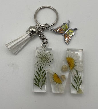A metal keychain featuring a white tassel, a butterfly charm, and two rectangular clear resin letters spelling 'M', each containing pressed white daisies with yellow centers and green stems.