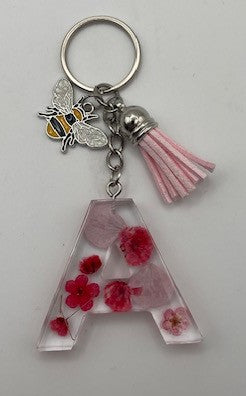 A silver metal keychain featuring a large clear resin letter 'A' filled with pink and red pressed flowers, a silver bee charm, and a pink tassel.