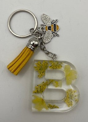 A decorative keychain featuring a silver metal key ring, a yellow tassel, a bee charm, and a clear resin letter 'B' filled with yellow dried flowers, shown on a plain light background.