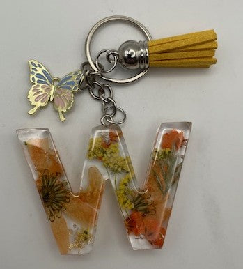 A letter 'W' keychain pendant made of clear resin with dried orange flowers and yellow botanicals, attached to a silver key ring with a gold butterfly charm and a yellow tassel.