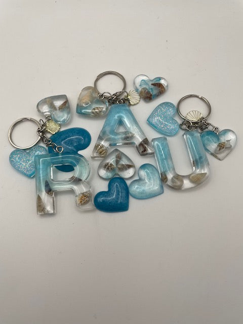 A set of personalized keychains featuring the name 'JUDITH' spelled out in translucent blue and clear acrylic letters, decorated with seashell charms and blue glitter hearts, on a plain light background.