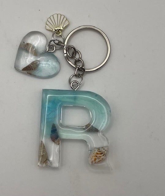 A personalized keychain featuring a large resin letter 'R' and a matching heart charm, both filled with small seashells and decorated with light blue and white resin, attached to a silver metal key ring.