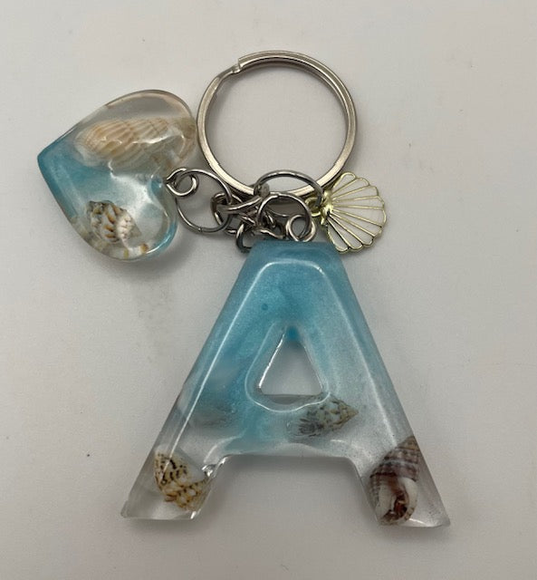 A resin letter 'A' keychain with embedded seashells and a matching heart-shaped charm on a silver keyring, photographed on a plain light background.