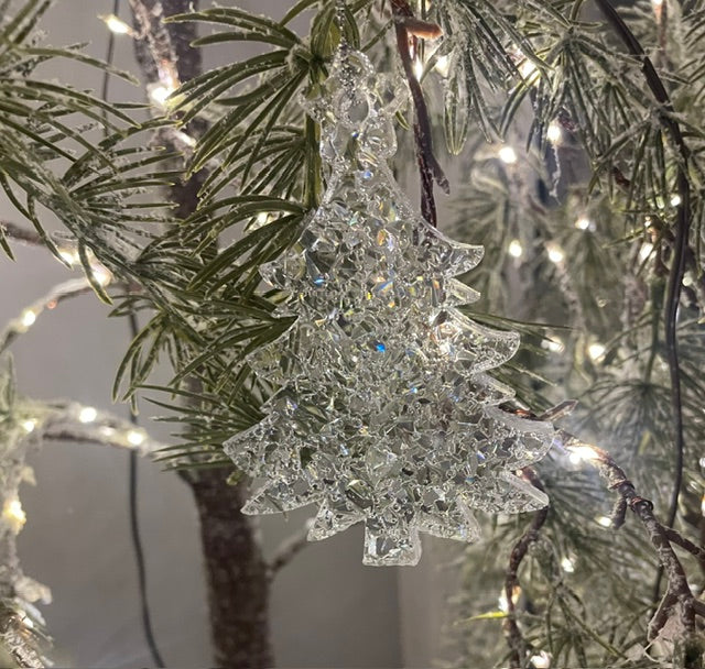 A clear, faceted tree-shaped Christmas ornament hanging from a pine tree branch with warm white string lights in the background.