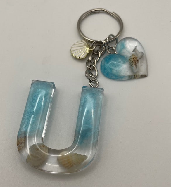 A handmade resin keychain featuring a large letter 'U' charm filled with light blue resin and seashells, a small heart charm with a seashell inset, and a gold seashell accent, all attached to a silver metal keyring.