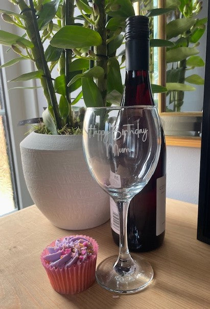 Wine glass with 'Happy Birthday Mum' engraving, bottle of wine, and cupcake on a table.