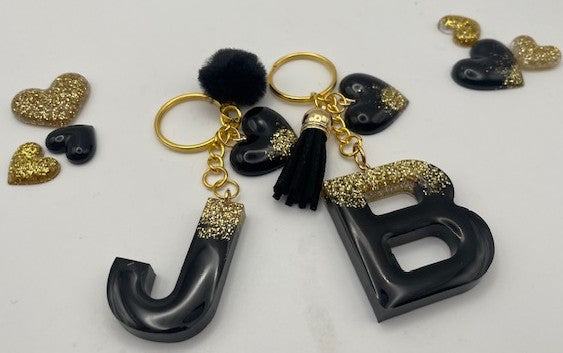 A set of personalized keychains featuring glossy black resin letters 'J' and 'B' with gold glitter accents, black tassel charms, and small black heart charms with gold glitter