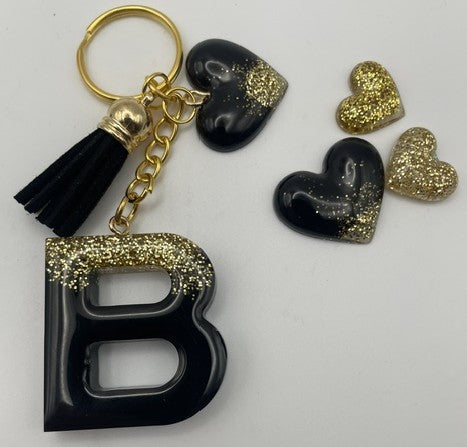 A black and gold letter 'B' keychain with a black tassel, shown alongside three matching heart-shaped charms on a plain white background.