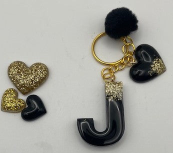 A black and gold keychain featuring a glossy black letter 'J' charm, a black heart charm with gold glitter, a black pom-pom, and gold-tone rings, displayed alongside three small heart charms on a white background.
