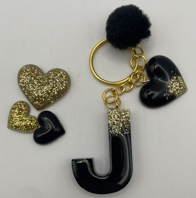 A black and gold keychain featuring a glossy letter 'J' charm, a black pom-pom, a gold ring, and three heart charms in black and glittery gold, displayed on a white background.