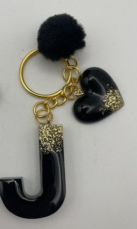 A personalized keychain featuring a black letter 'J' charm, a black heart charm with gold glitter, a black pom-pom, and gold-tone metal hardware, shown on a plain light background.