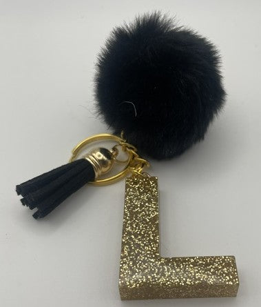 A personalized keychain featuring a black faux fur pom-pom, a black tassel with a gold cap, a gold split key ring, and a glittery gold letter 'L' charm, displayed on a light gray background.
