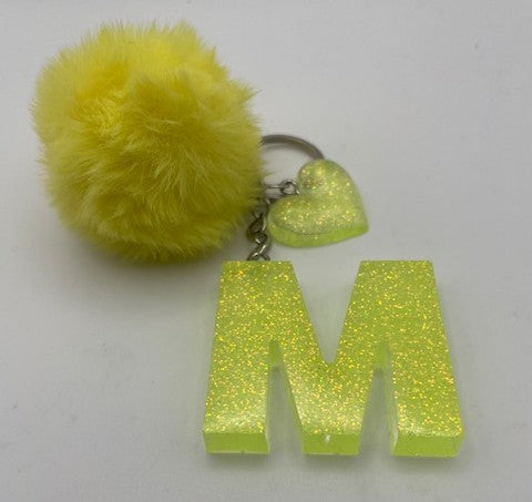 A neon yellow glitter monogram keychain featuring a large letter 'M' charm, a small glitter heart charm, and a fluffy yellow pom-pom on a silver key ring, set against a plain white background.