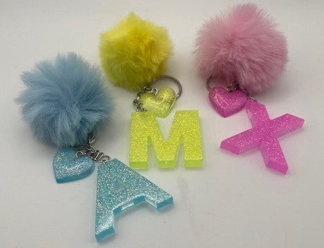 Three personalized acrylic letter keychains with fluffy pom-poms, displayed on a white background. The keychains feature glittery letters 'M' and 'A' in yellow and blue, alongside matching glittery heart charms and pom-poms in yellow, pink, and light blue.