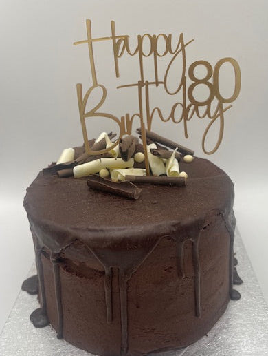 Chocolate cake with decorative elements and a 'Happy 80th Birthday' topper on a light gray background.