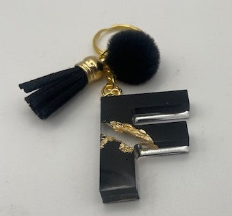 A black resin letter 'F' keychain with gold leaf detailing, a black faux fur pom pom, a black tassel, and gold-tone metal hardware, shown on a light gray background.
