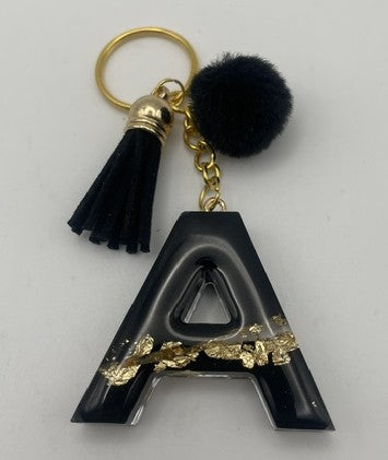 A black resin letter 'A' keychain with gold foil accents, a black tassel, a black pom-pom, and gold-tone hardware on a light gray background.