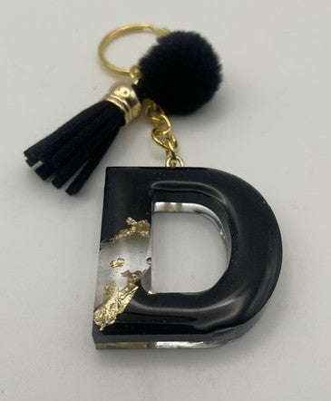 A black resin letter 'D' keychain charm with gold foil accents, a black pom-pom, and a black tassel on a gold ring, shown on a light gray background.