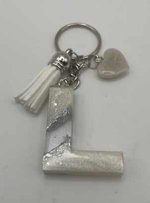 A silver-toned metal keychain featuring a large textured letter 'L' charm, a white tassel, and a small heart-shaped charm, shown on a plain light background.