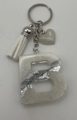 A silver-toned metal keychain featuring a large glittery letter 'B' charm, a small white tassel, a small white heart-shaped charm, and a silver ring, shown on a plain light background.