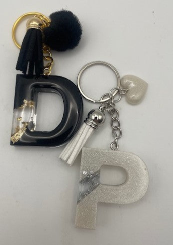 A personalized keychain featuring two resin letters, 'D' in black with gold foil flakes and a black pom-pom tassel, and 'P' in glittery silver with a small silver tassel. The keychain also includes a silver key ring, a small white heart charm, and a white tassel.