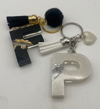 A personalized keychain featuring a glittery silver letter 'P' charm, a black tassel, a black rectangular plaque with gold leaf accents, a white tassel, a black pom-pom, and a small white heart charm on a silver keyring.