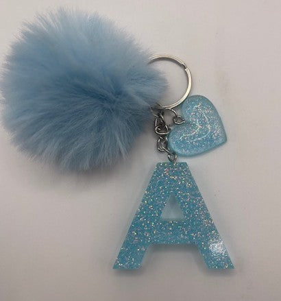A light blue glittery keychain featuring a large fuzzy pom-pom, a silver split key ring, a small glittery heart charm, and a glittery letter 'A' pendant.